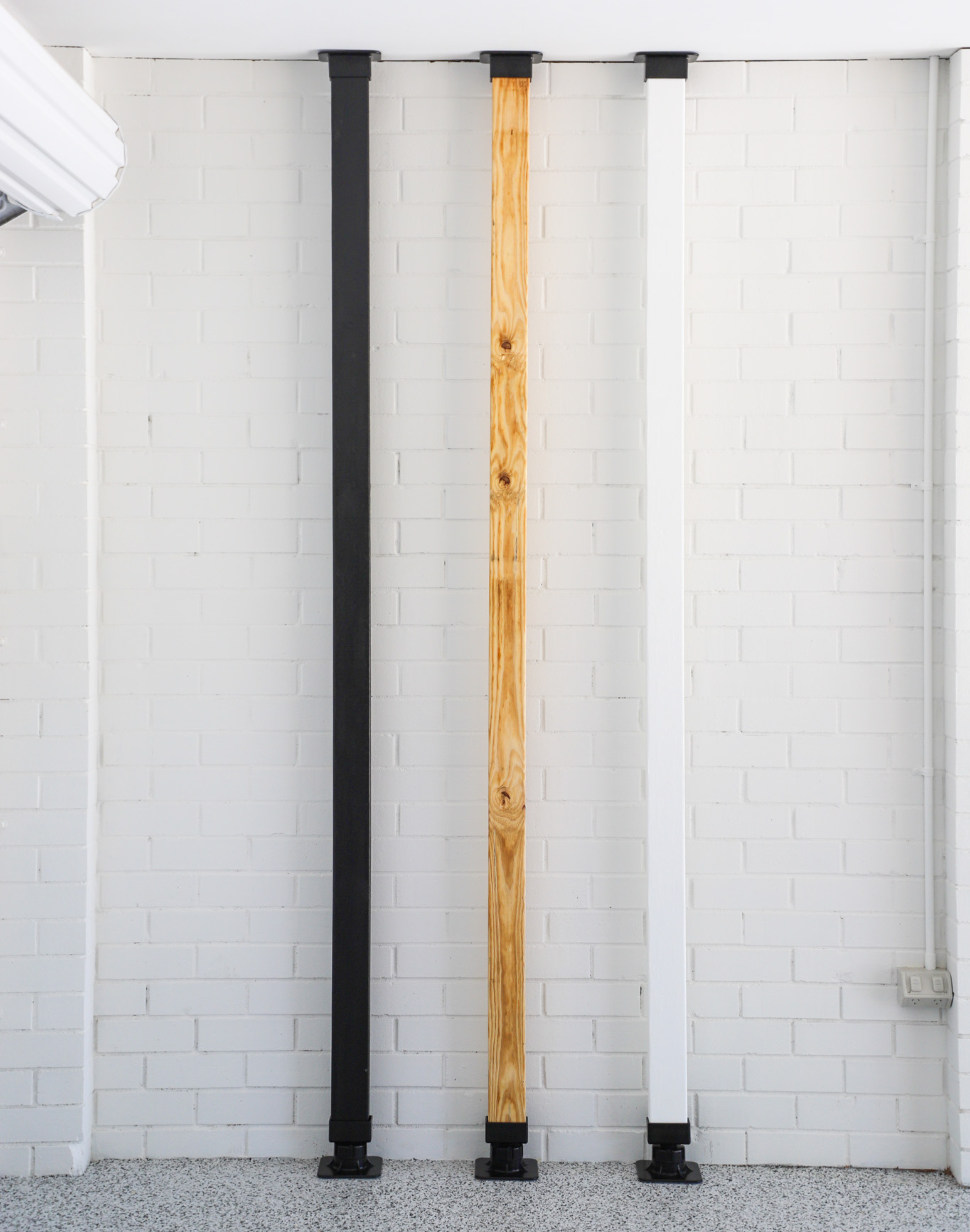 A white, raw and black wooden pole with black extendable bases on the top and bottom, on a white brick background. 